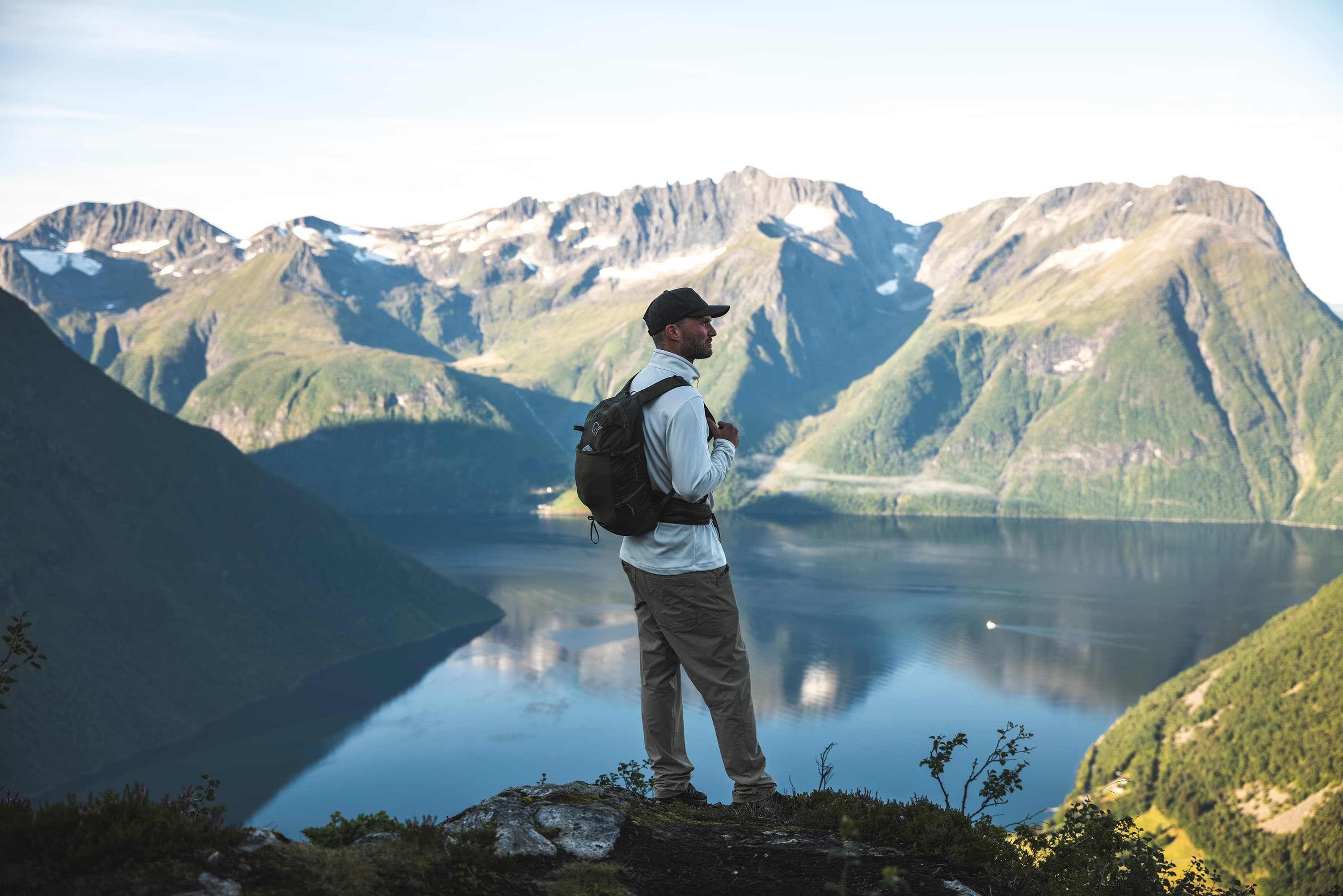 A person with a backpack stands on a cliff, overlooking a tranquil fjord surrounded by rugged mountains under a clear blue sky.