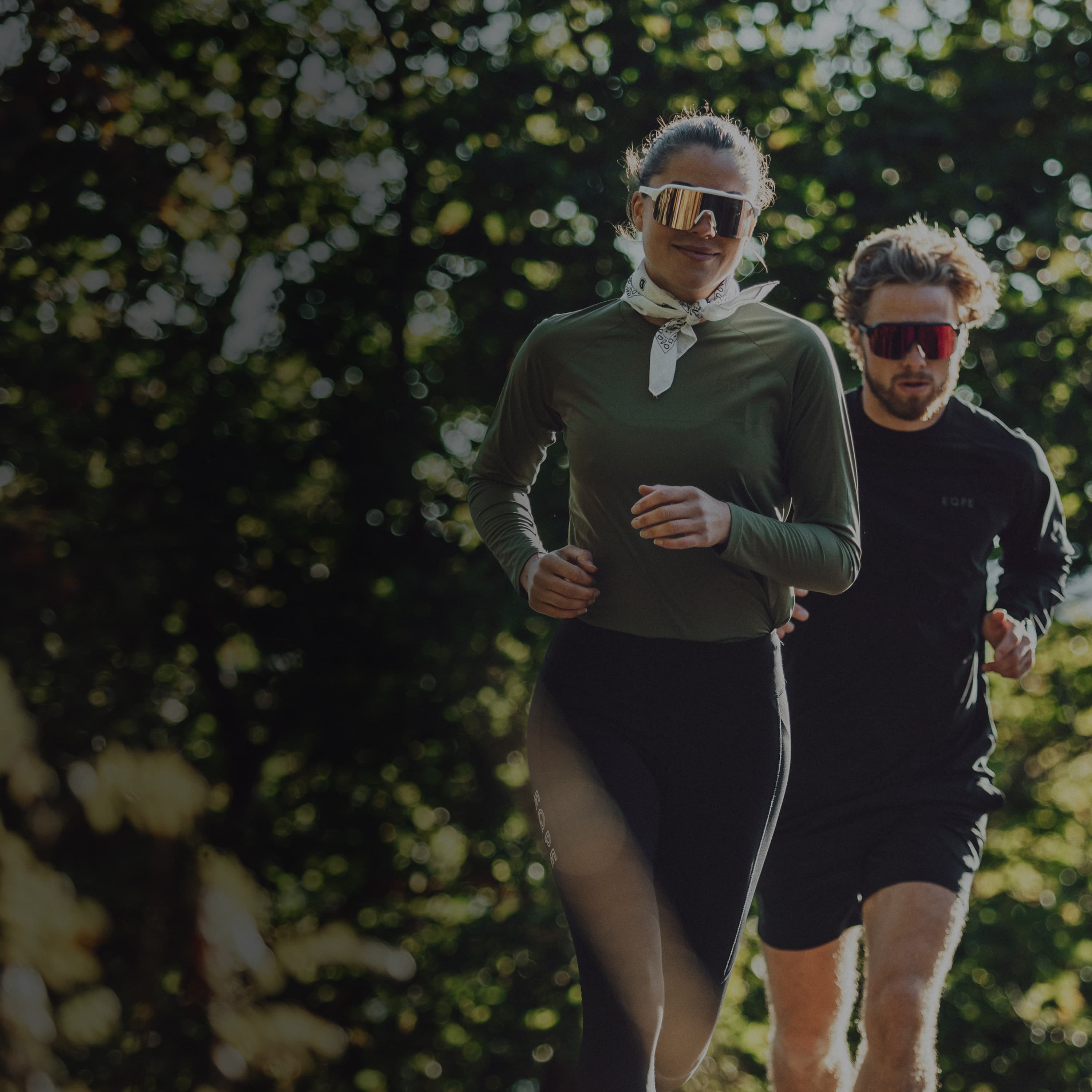 Two people jogging outdoors in a wooded area, wearing long sleeves and reflective sunglasses, surrounded by lush green foliage.