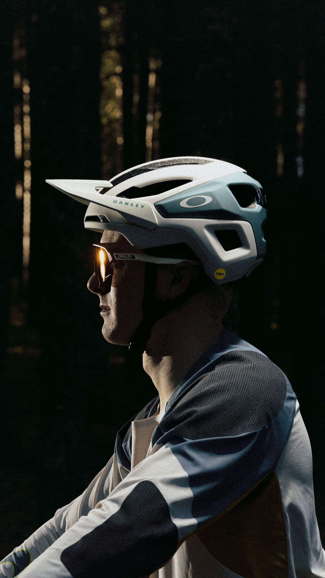 Cyclist wearing a helmet and sunglasses, captured in profile against a dark, forested background.