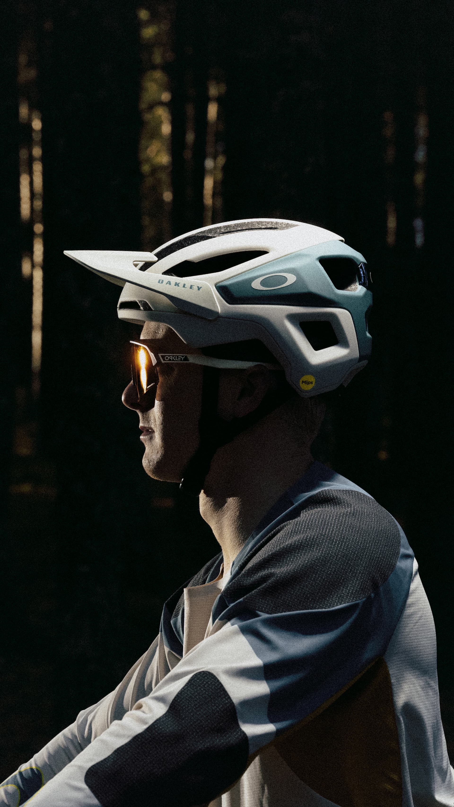 Cyclist wearing a helmet and sunglasses, captured in profile against a dark, forested background.