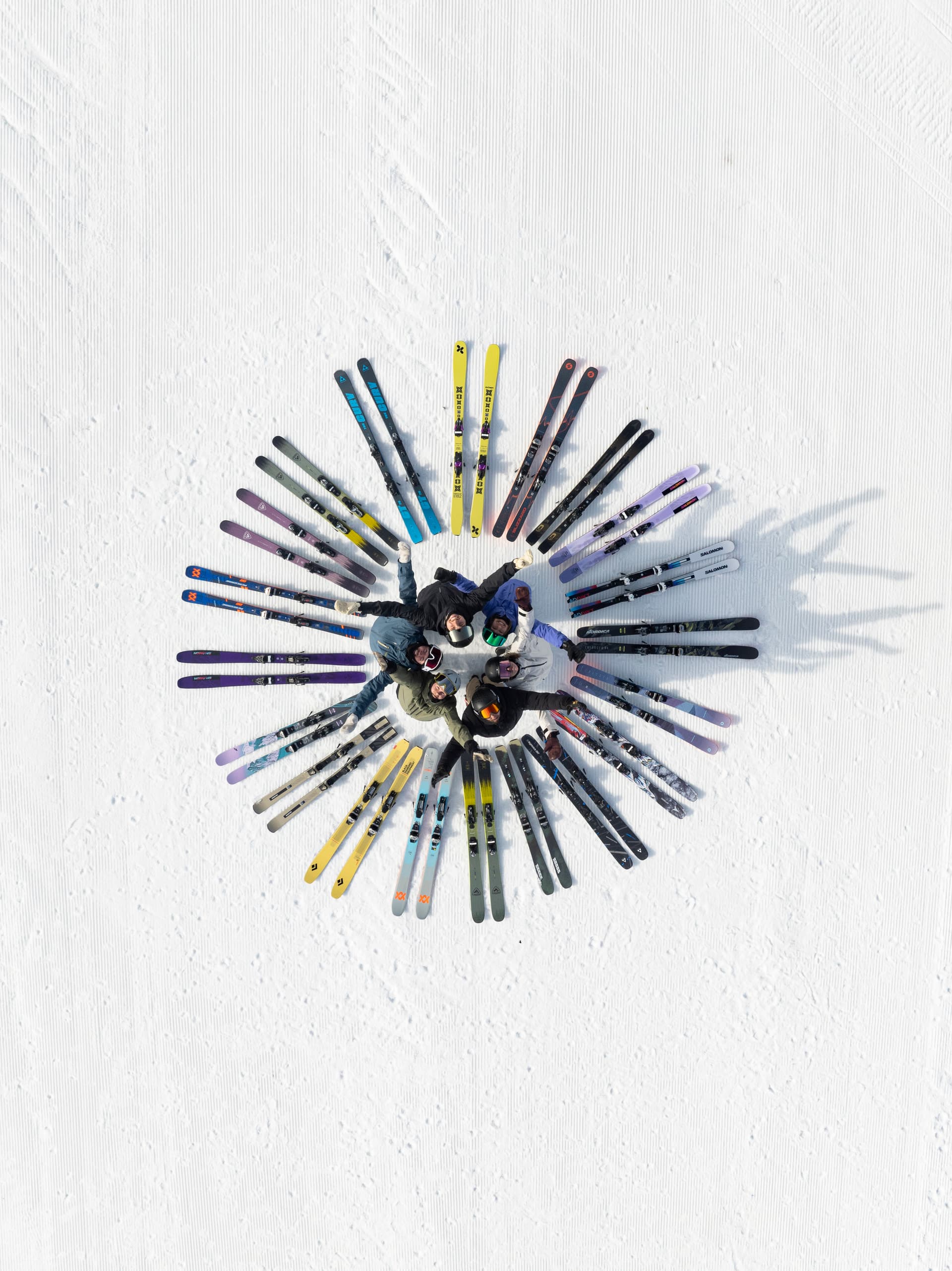 Aerial view of people standing in a circle on snow, heads together, surrounded by skis radiating outward like a sunburst pattern.