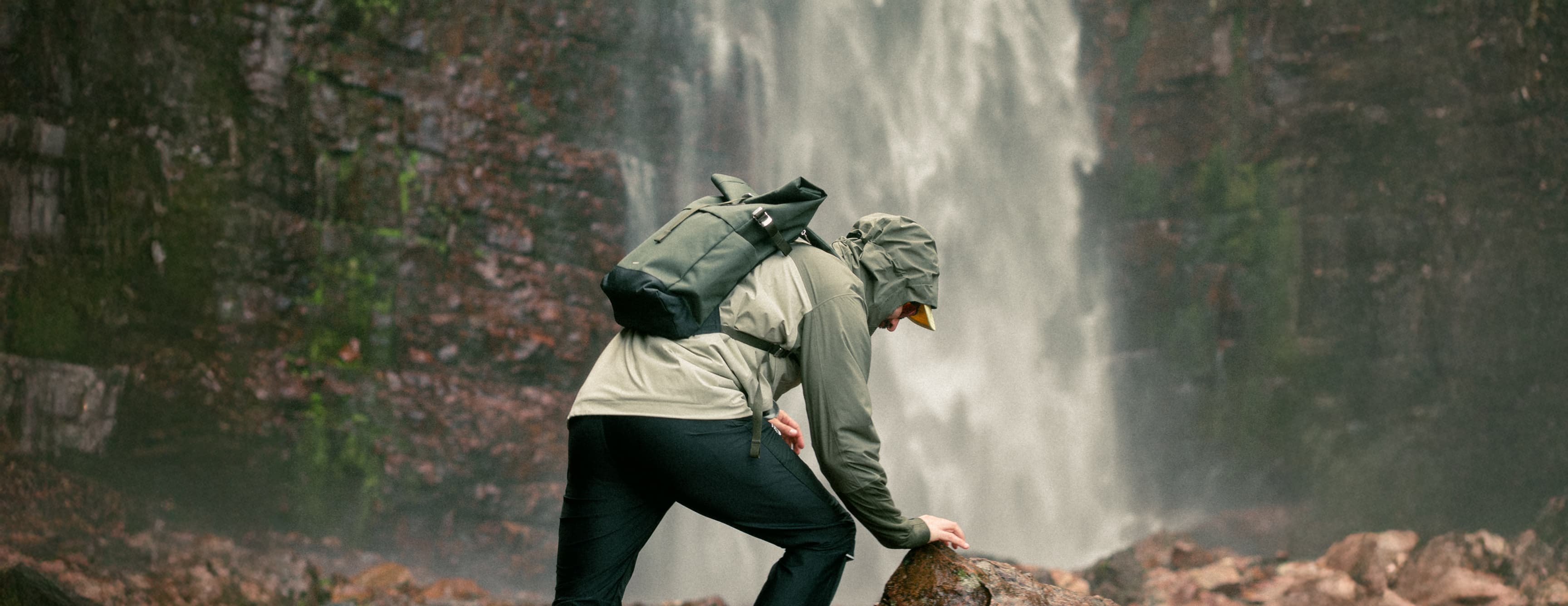 Person in outdoor gear climbing rocks near a waterfall, with mist and lush greenery in the background.