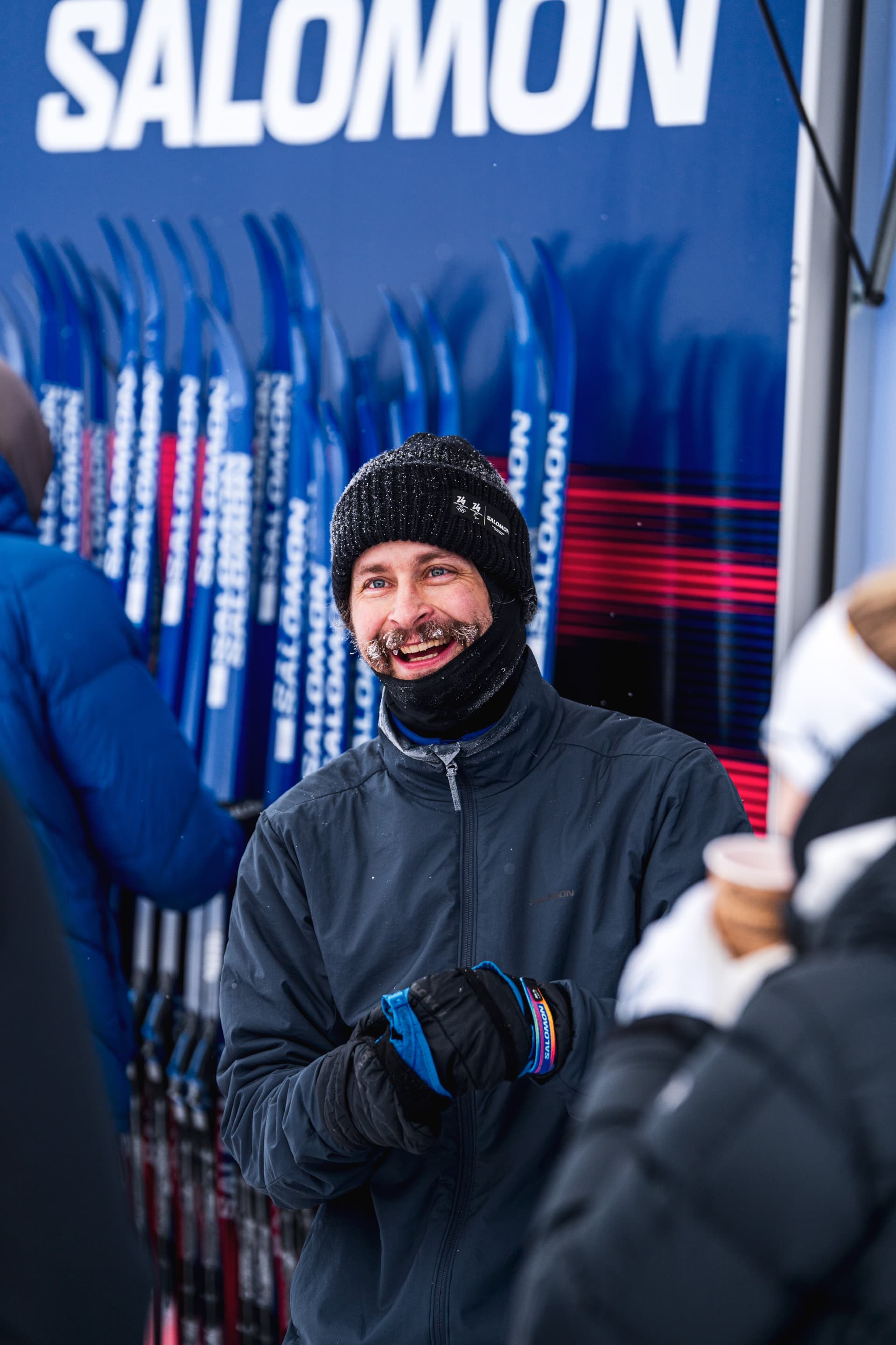 Man dressed warmly, smiling in front of rows of skis at a Salomon booth, with snowflakes falling around.