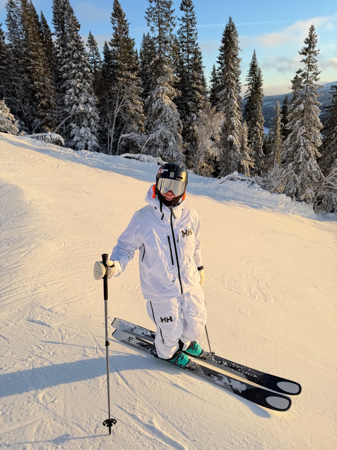 Skier Rasmus Stegfeldt in a white outfit and helmet stands on a snowy slope, surrounded by snow-covered trees under a clear sky.