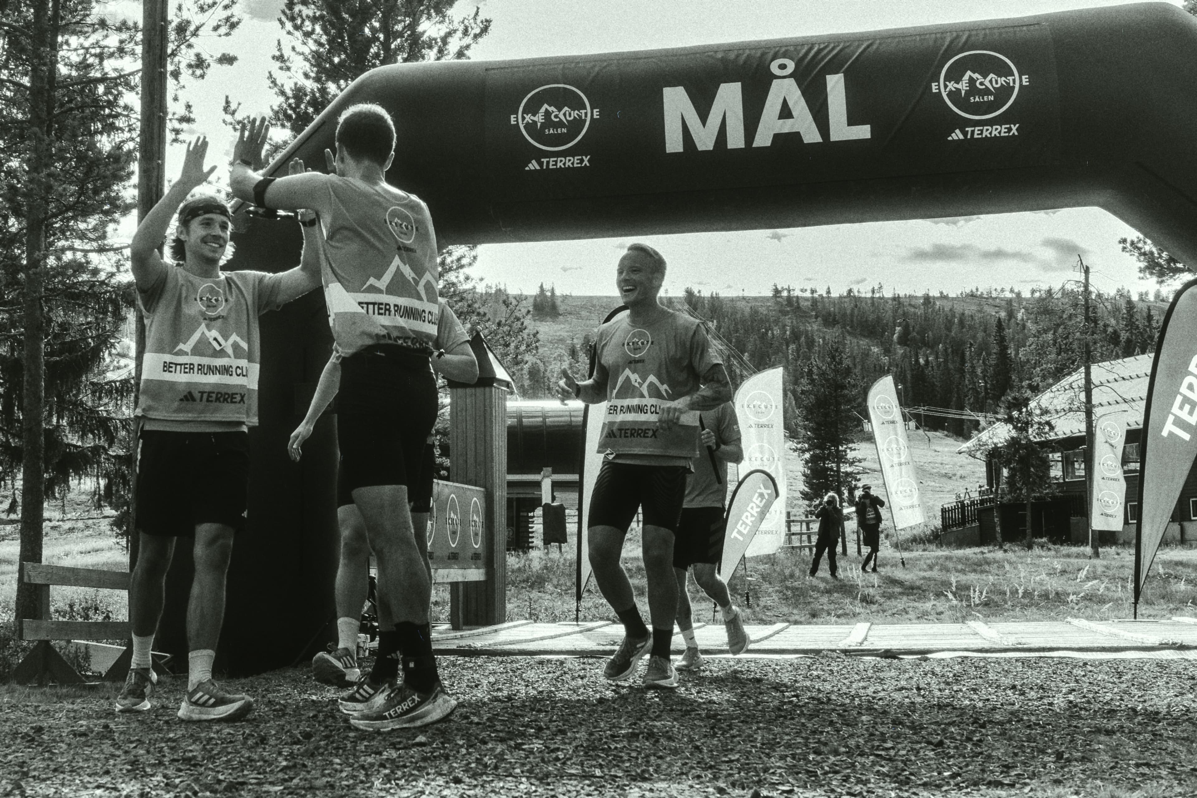Runners celebrating at the finish line, surrounded by trees and banners, in a black and white setting.
