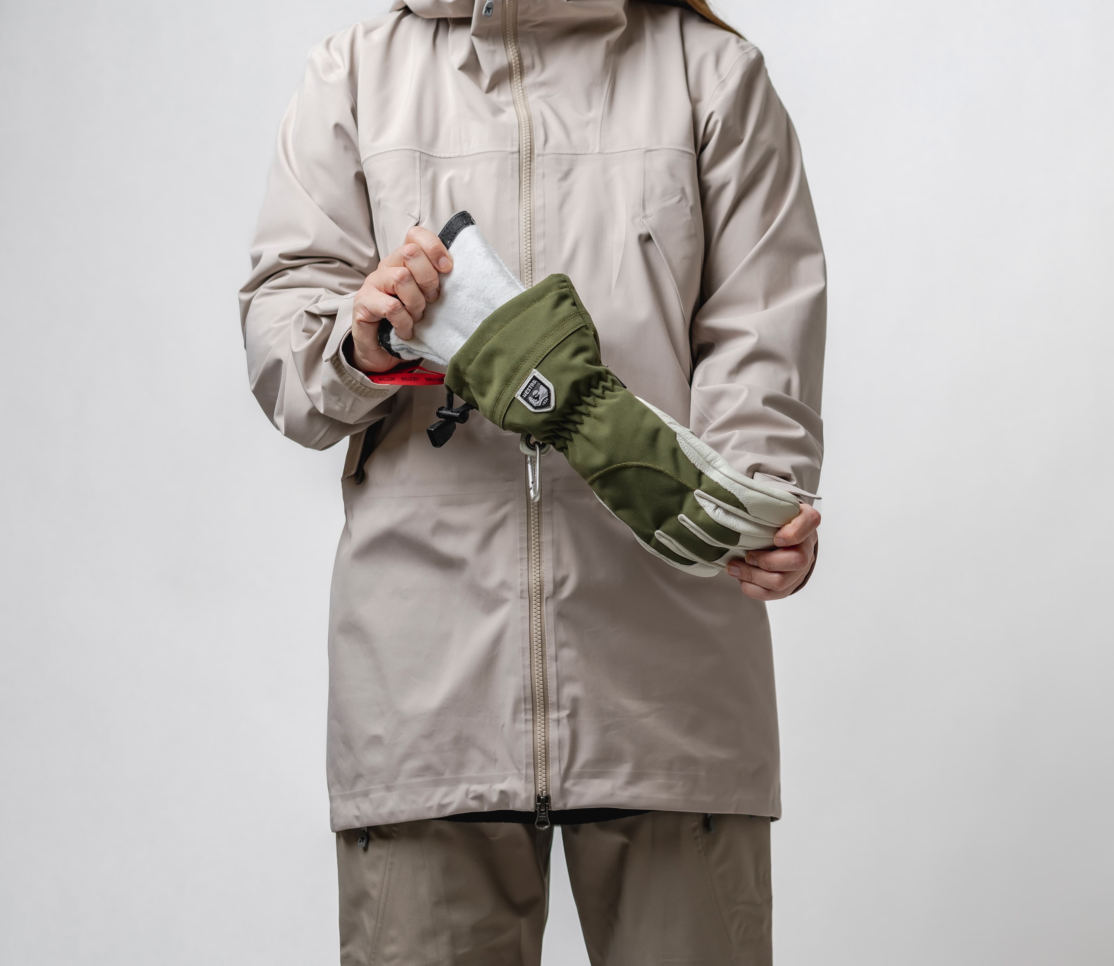 Person in a beige winter clothes holding a green and white glove against a plain background.