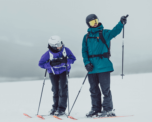 Two skiers in winter gear stand on a snowy slope, one pointing into the distance.