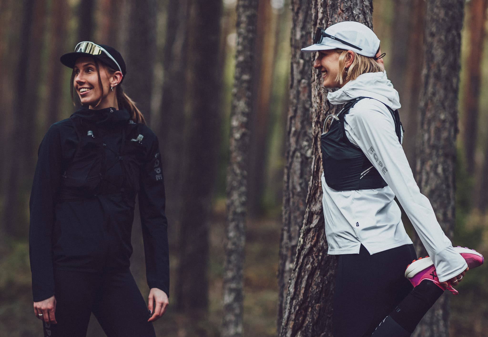 Two women in athletic gear stand in a forest. One stretches her leg while the other smiles, both wearing caps and vests.