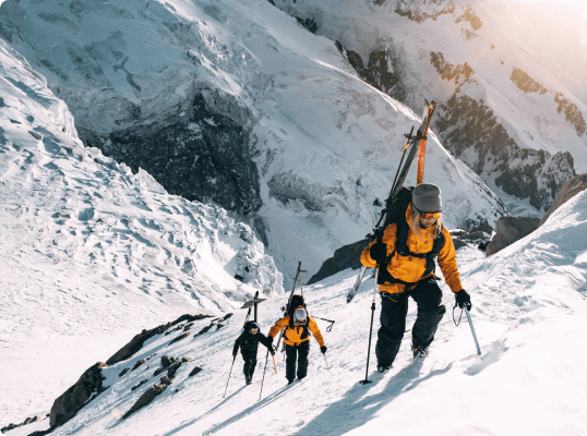 Three climbers in yellow jackets ascend a snowy, steep mountain slope with skis on their backs.