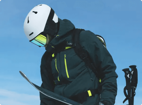 Skier in a green jacket and white helmet, holding skis against a clear blue sky.