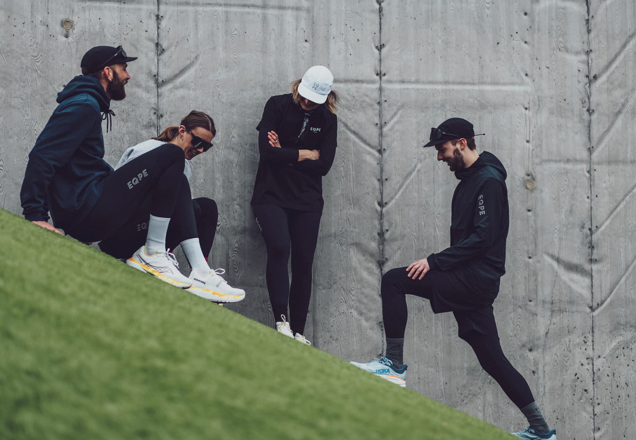 Four people in athletic wear on a grassy slope beside a concrete wall.
