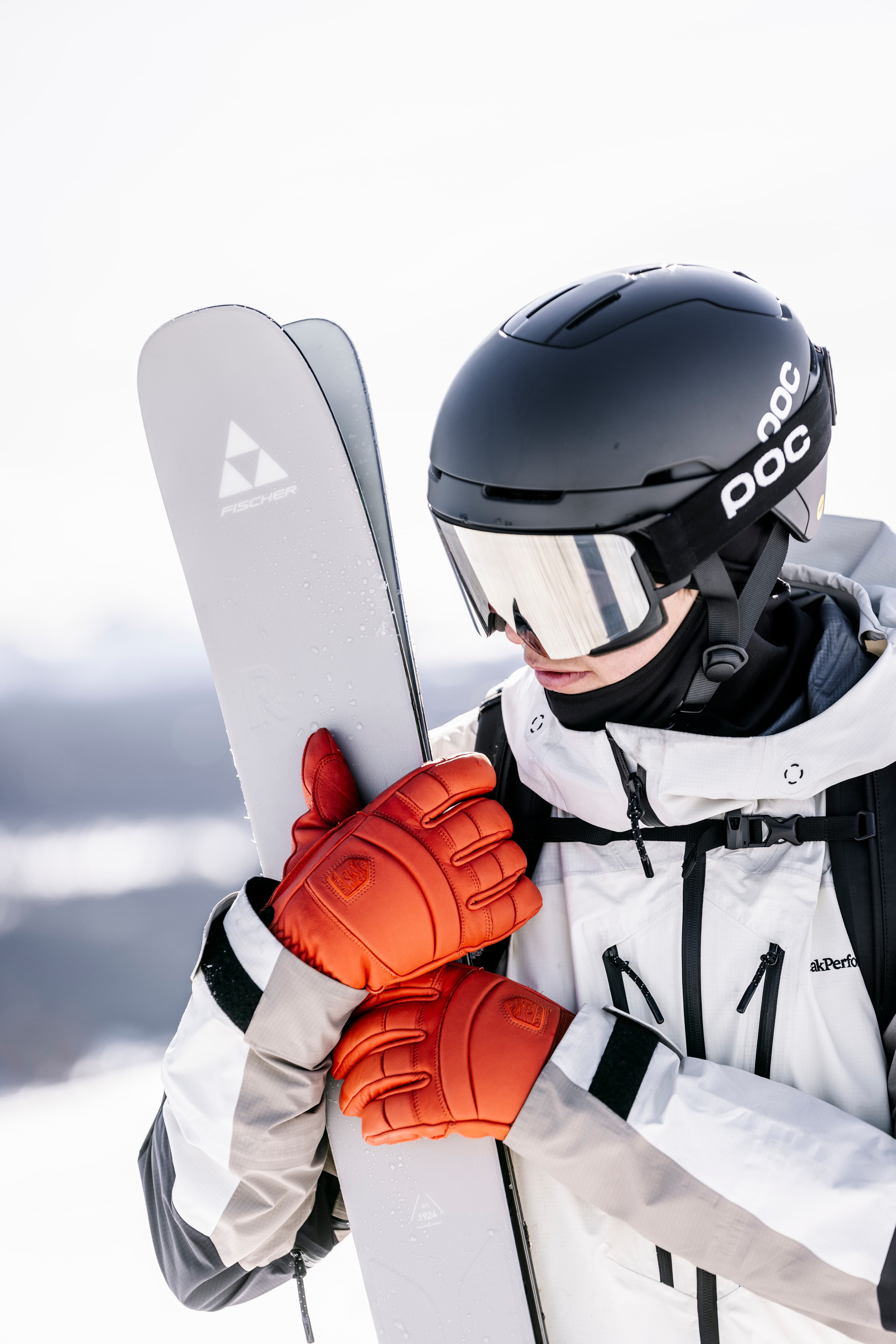 A skier in a white jacket and orange gloves holds a pair of skis in a snowy landscape.