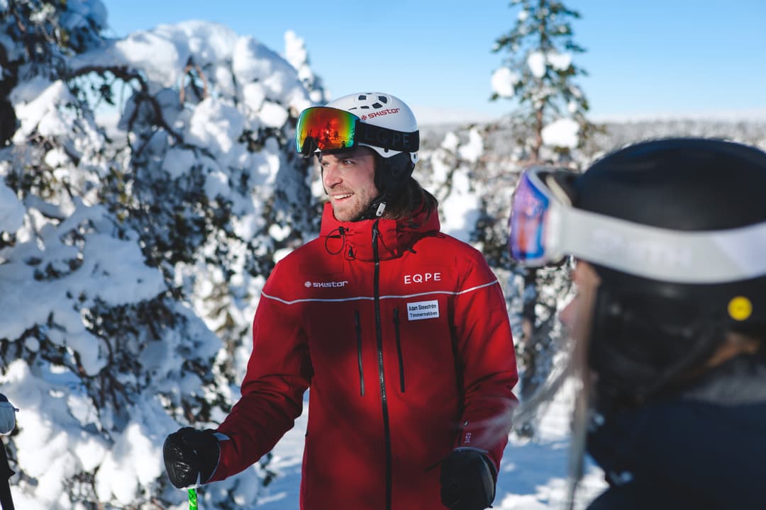 A person in a red jacket and ski helmet holds ski poles, standing in a snowy landscape with trees. Another person is partially visible.
