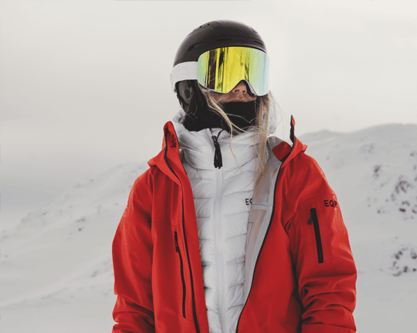Person in a red jacket and ski goggles stands in a snowy mountain landscape, looking into the distance.