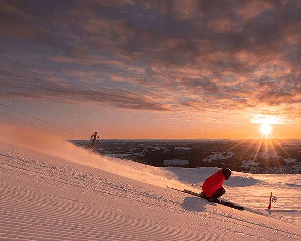 Skier in red jacket descends a snowy slope at sunset, with dramatic clouds and sun casting a warm glow over the landscape.