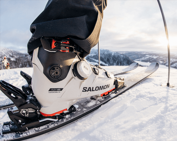 Close-up of a ski boot a snowy mountain slope, with distant snow-covered hills.