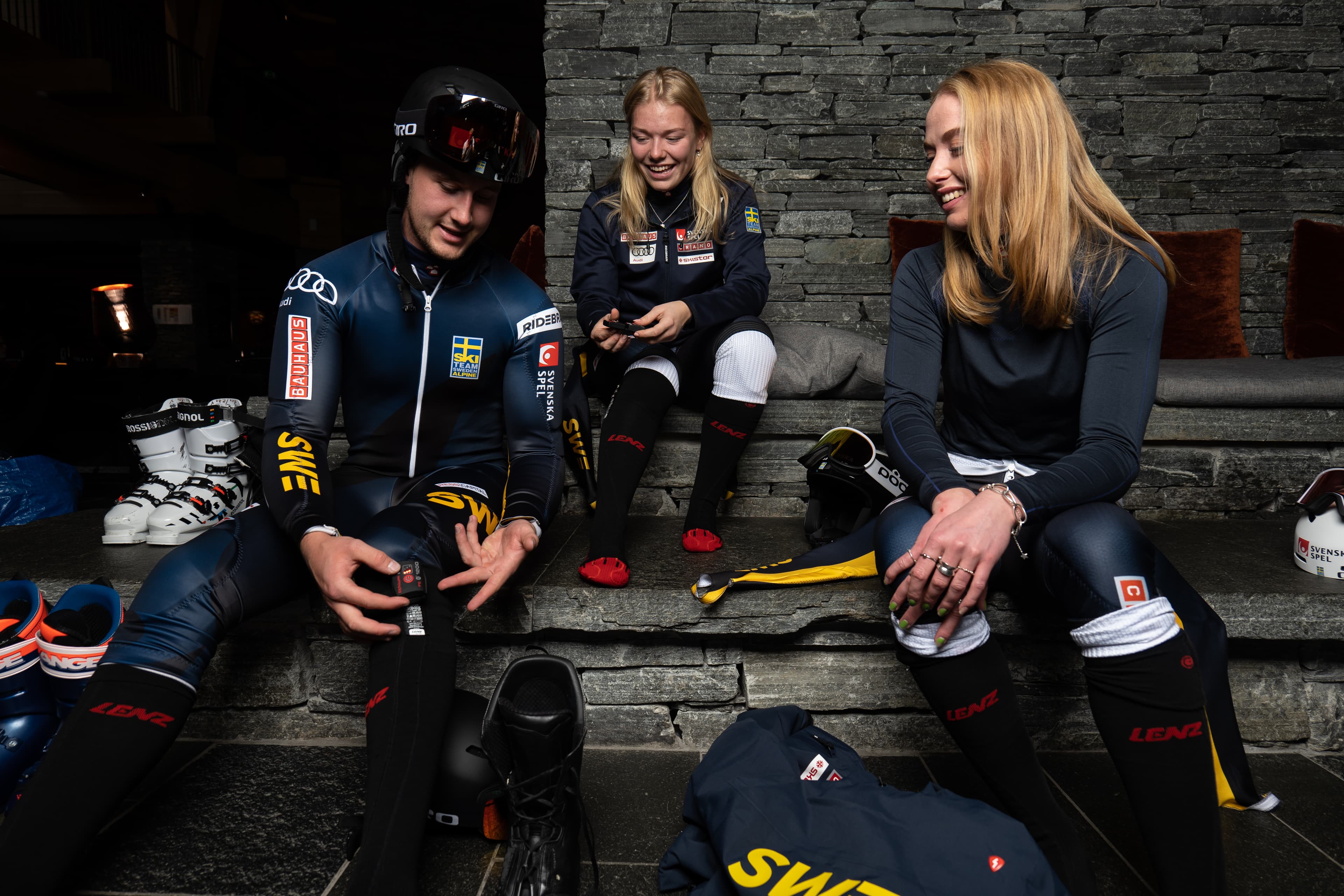 Three athletes in ski suits sit on a stone bench, chatting and preparing gear, with helmets and boots nearby.