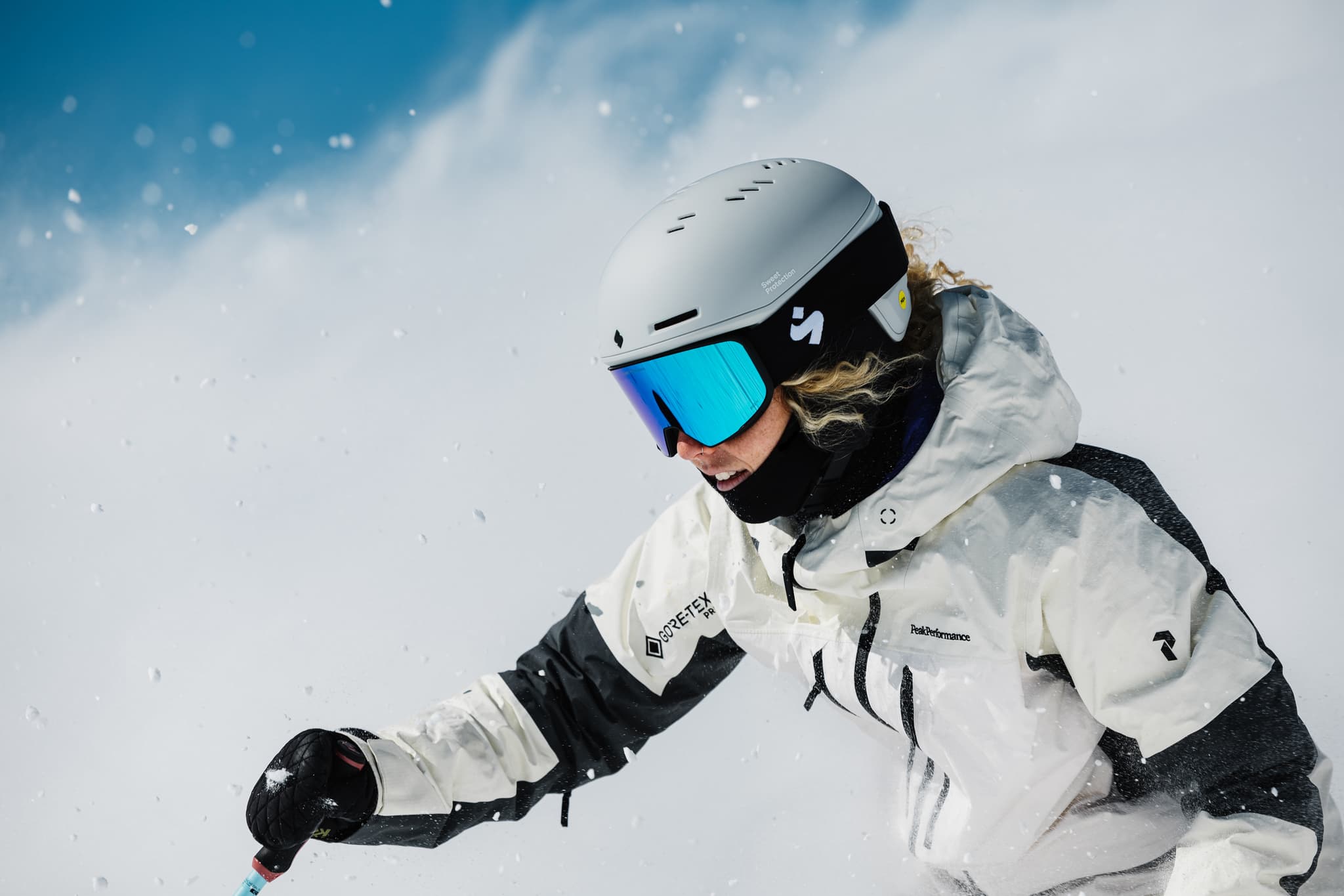 A skier in a white and black outfit and reflective goggles, skis down a snowy slope against a clear blue sky.
