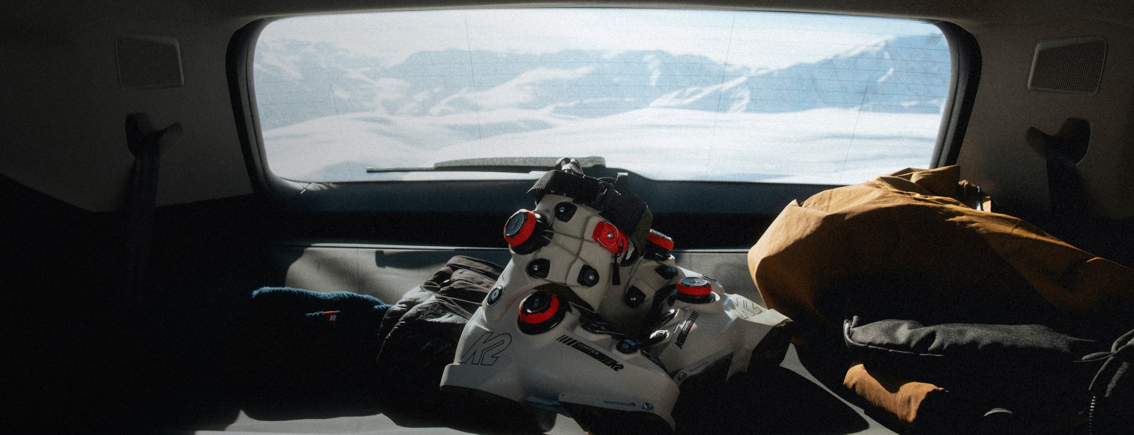 Ski boots and gear in a car trunk with a snowy mountain landscape visible through the rear window.