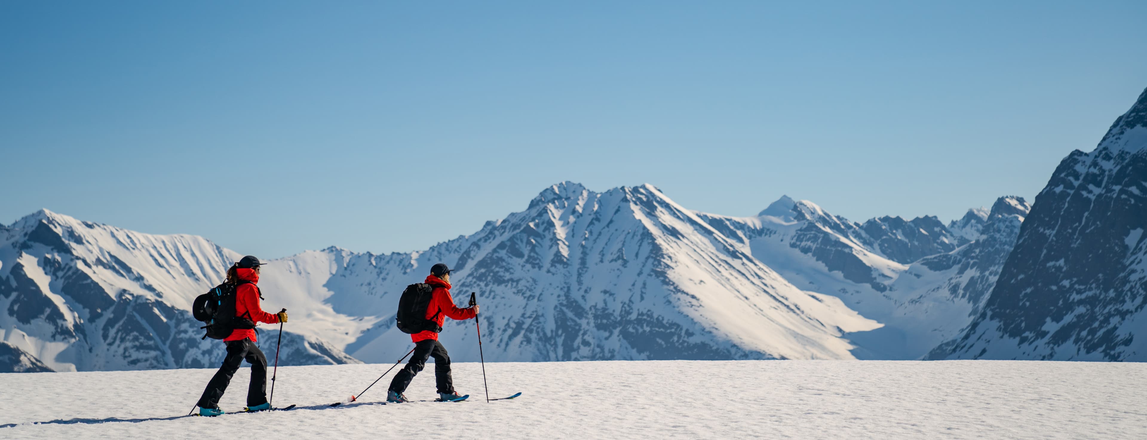 Two skiers in red jackets traverse a snowy mountain landscape under a clear blue sky, with rugged peaks in the background.