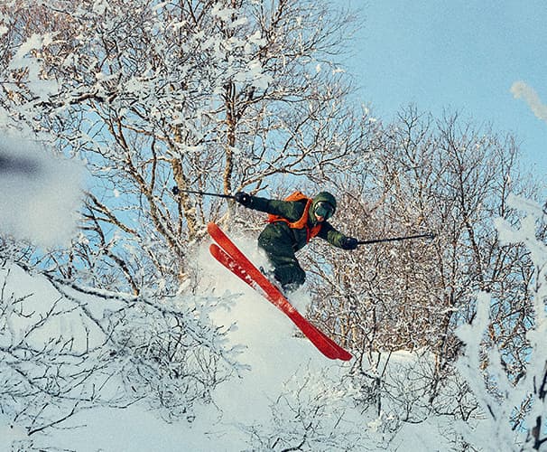 Skier in mid-air with red skis, surrounded by snow-covered trees.