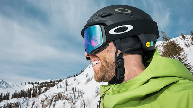 A person in a green jacket and black helmet smiles, with snowy mountains and a blue sky in the background.