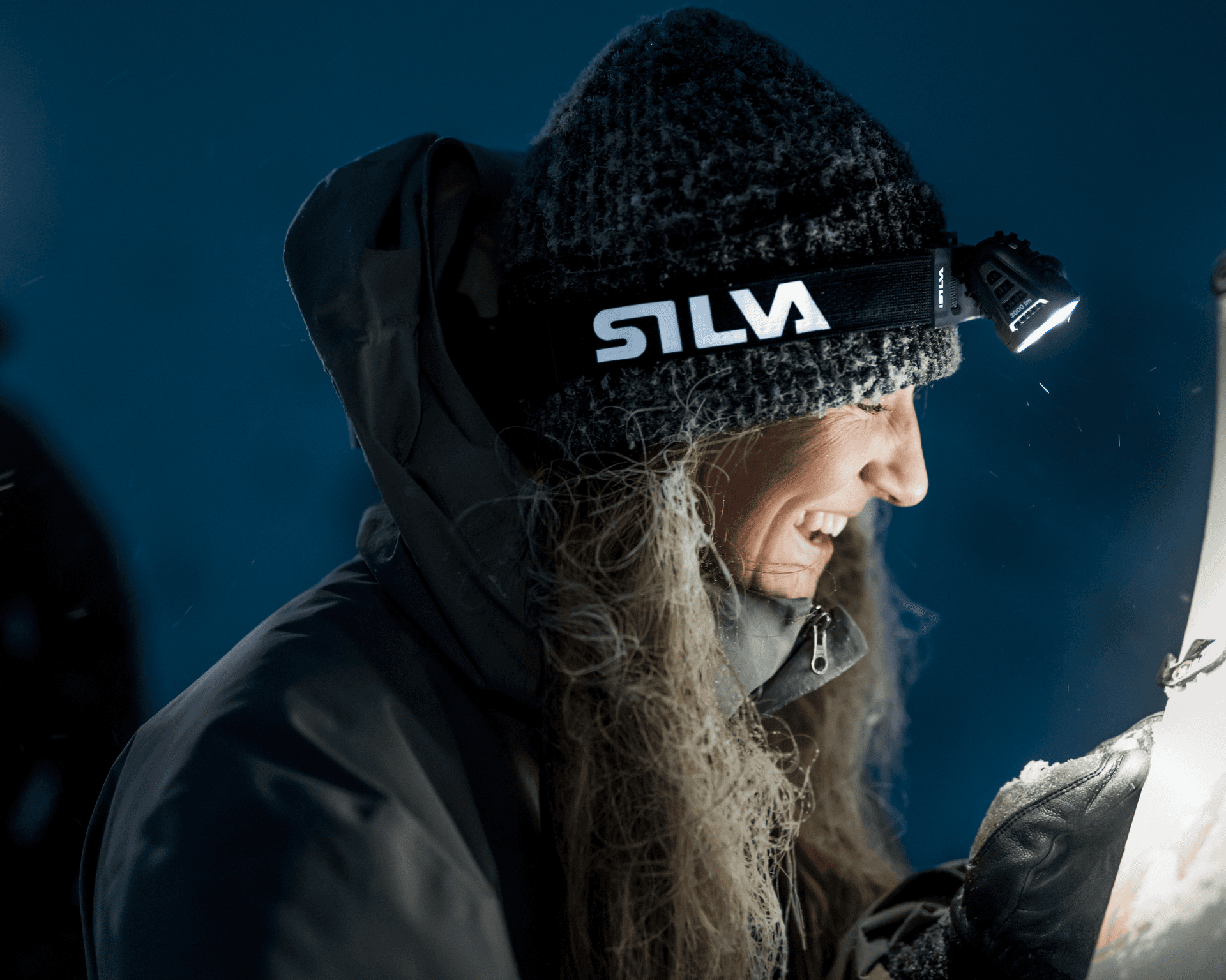 Smiling person, wearing a headlamp and winter clothing, in a dimly lit environment.