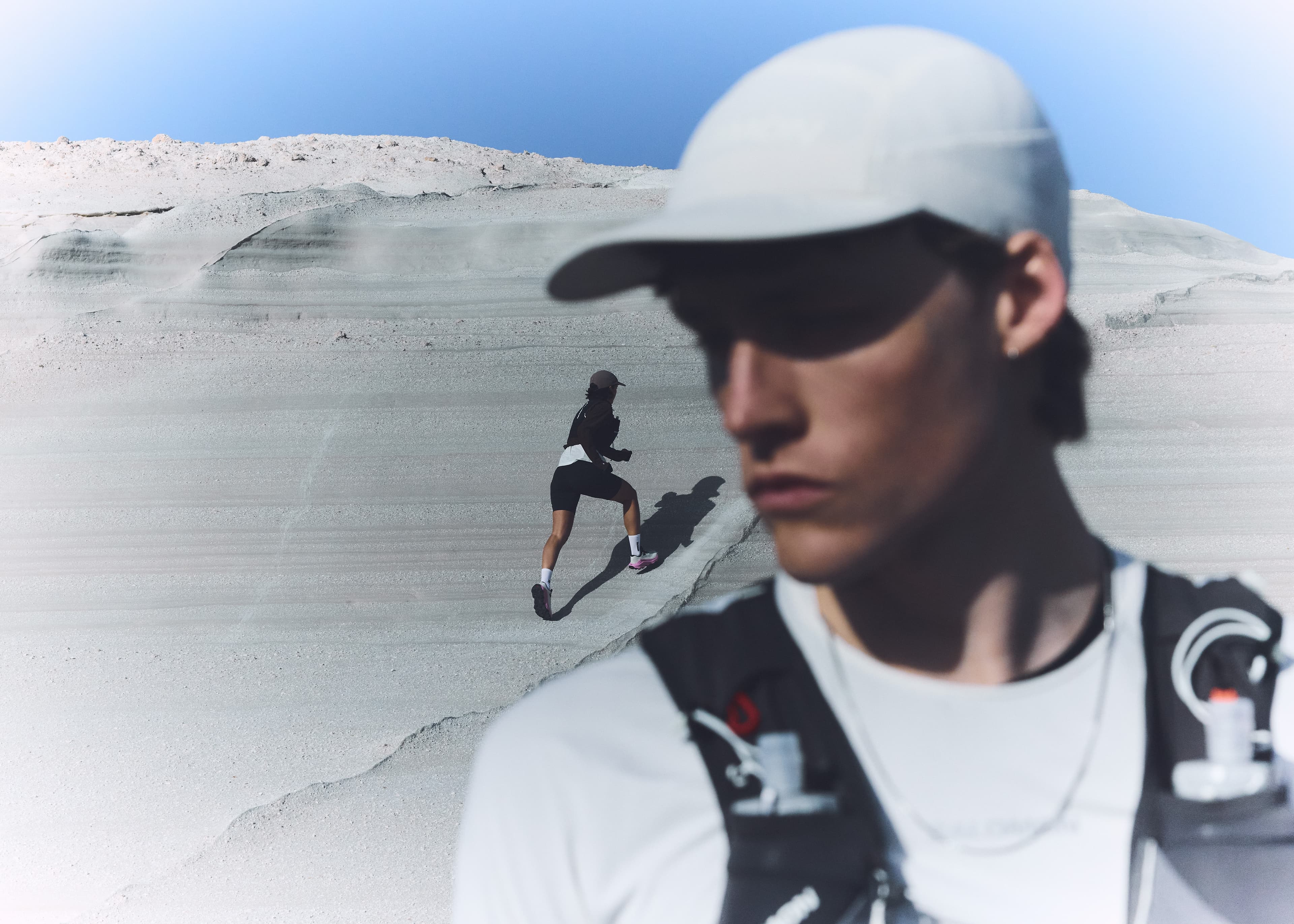 Two athletes in white caps and sports gear on a sandy slope; one in focus close-up, the other climbing in the background.