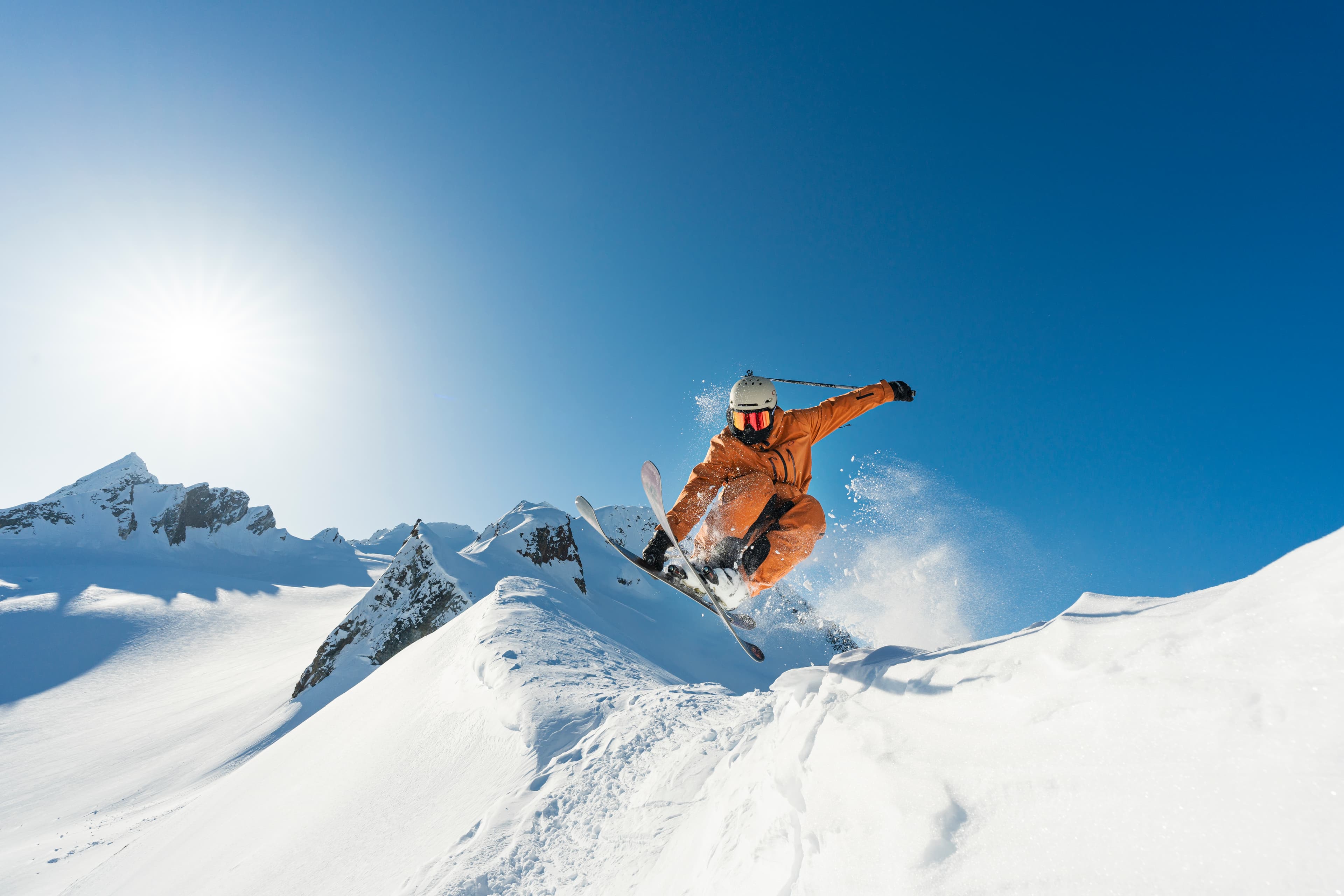 Skier in orange gear jumps off a snowy ridge against a clear blue sky, with sun shining brightly over snow-covered mountains.