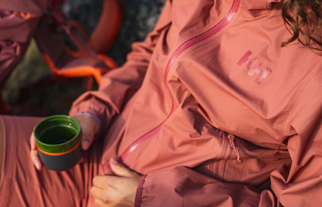 Person in a pink jacket holding a green cup, sitting outdoors with an orange backpack in the background.
