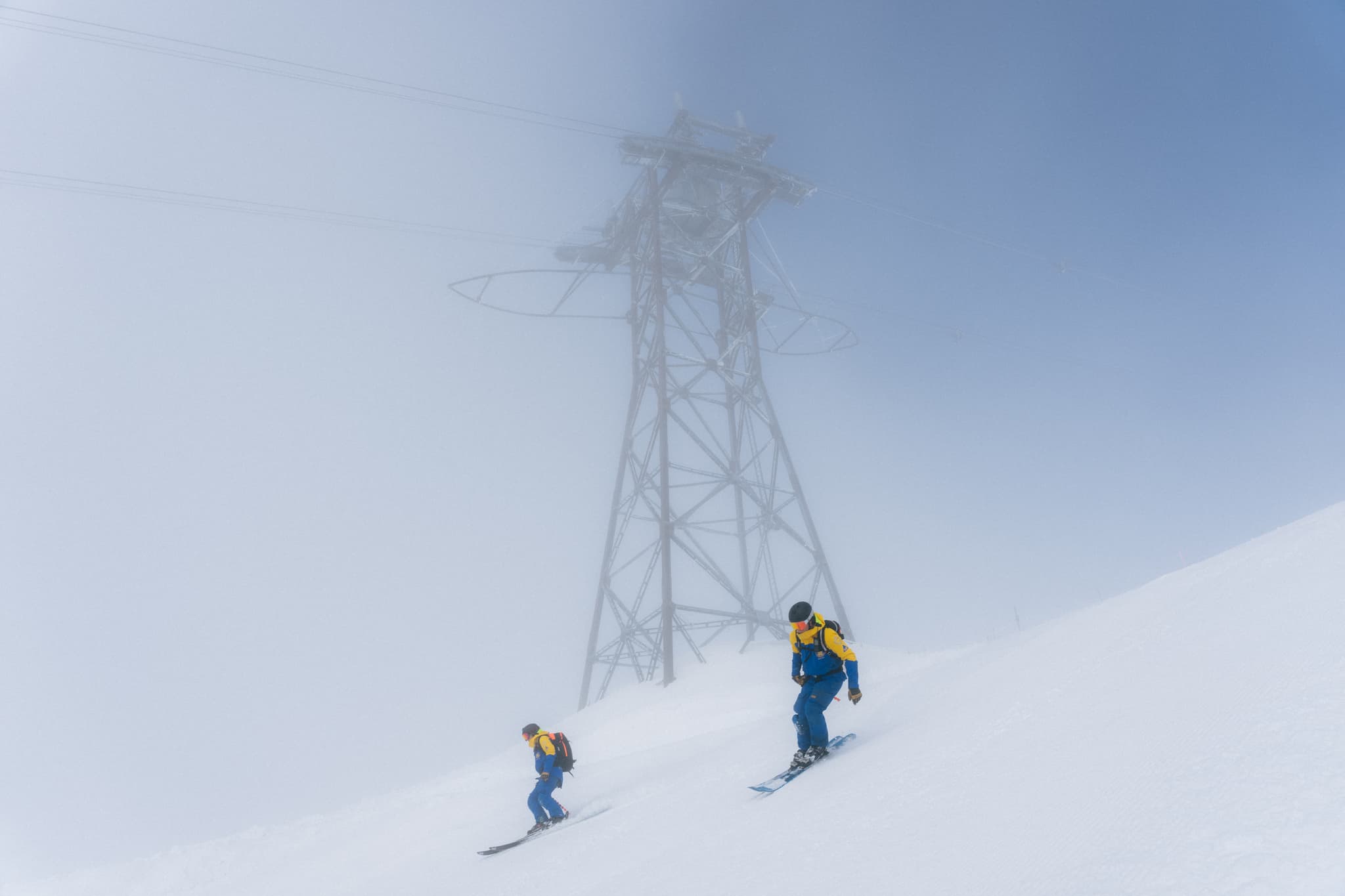 Two skiers in blue and yellow gear descend a snowy slope near a foggy metal tower on a misty day.