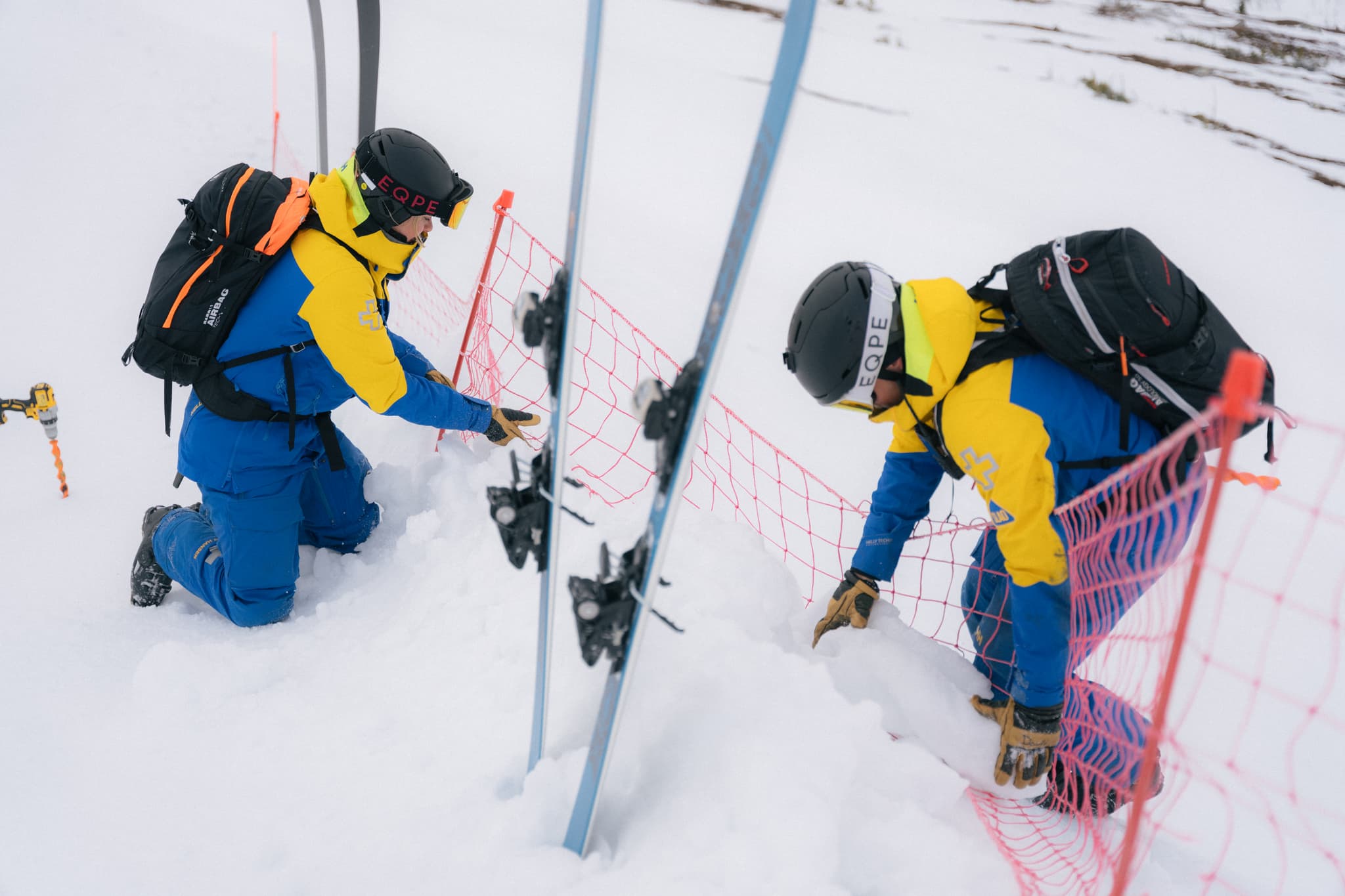 Two people in blue and yellow outfits work on a snowy slope, adjusting a red safety net.