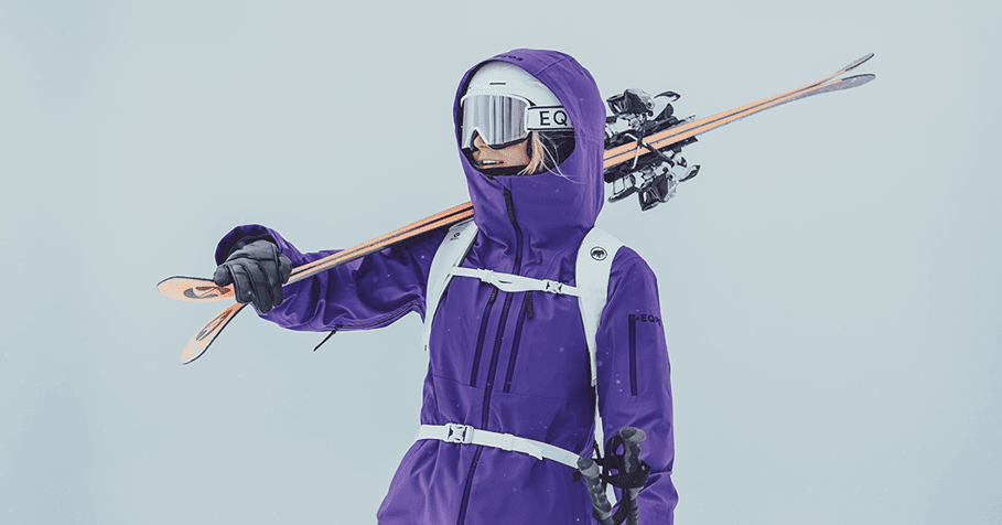 A person in a purple ski jacket and goggles carries skis on their shoulder.