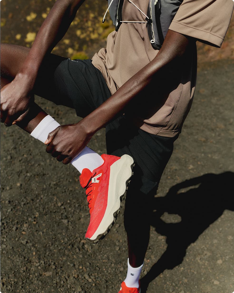 Person outdoors, wearing a brown shirt and black shorts, tying red sports shoes with white soles, and wearing a hydration vest.