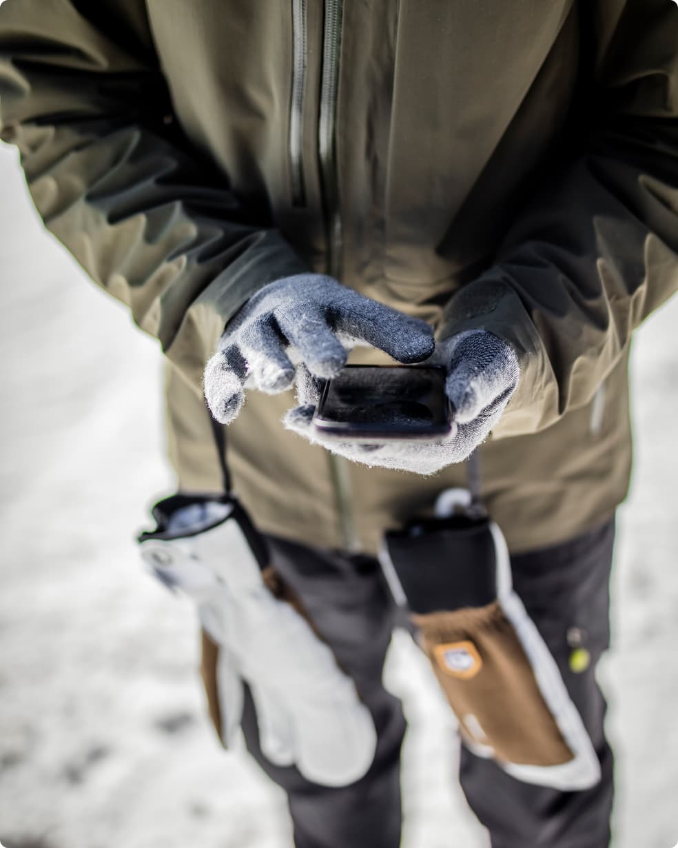 Person in winter clothing using a smartphone, wearing gray gloves, with hanging gloves on sides, standing in a snowy environment.