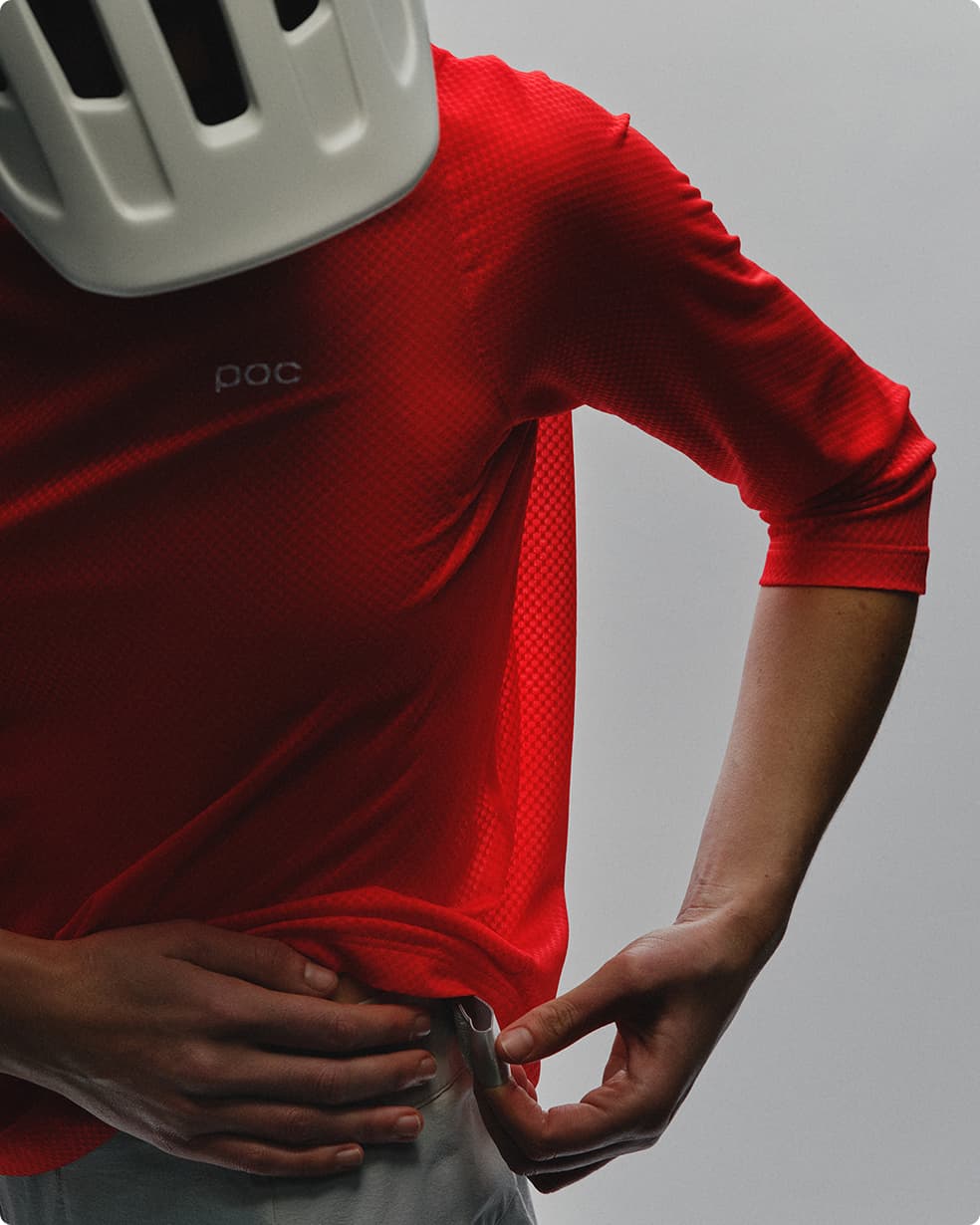 Person in a red cycling jersey adjusts the hem, wearing a white helmet against a neutral background.