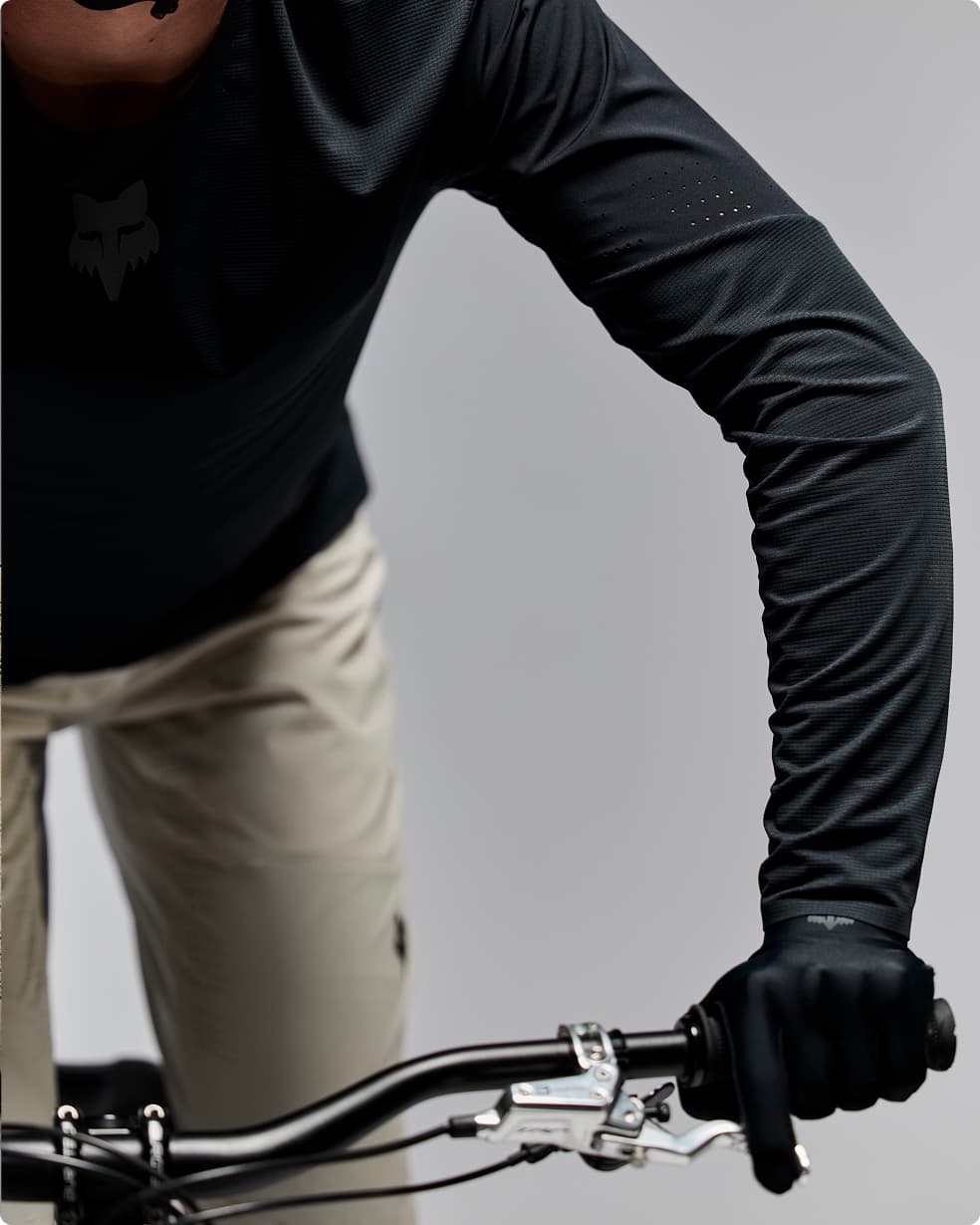 Close-up of a person in dark cycling gear and gloves holding mountain bike handlebars, wearing beige pants.