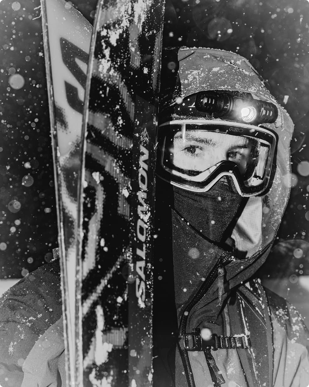 A skier in a snowstorm wearing a helmet, goggles, and headlamp stands beside Salomon skis