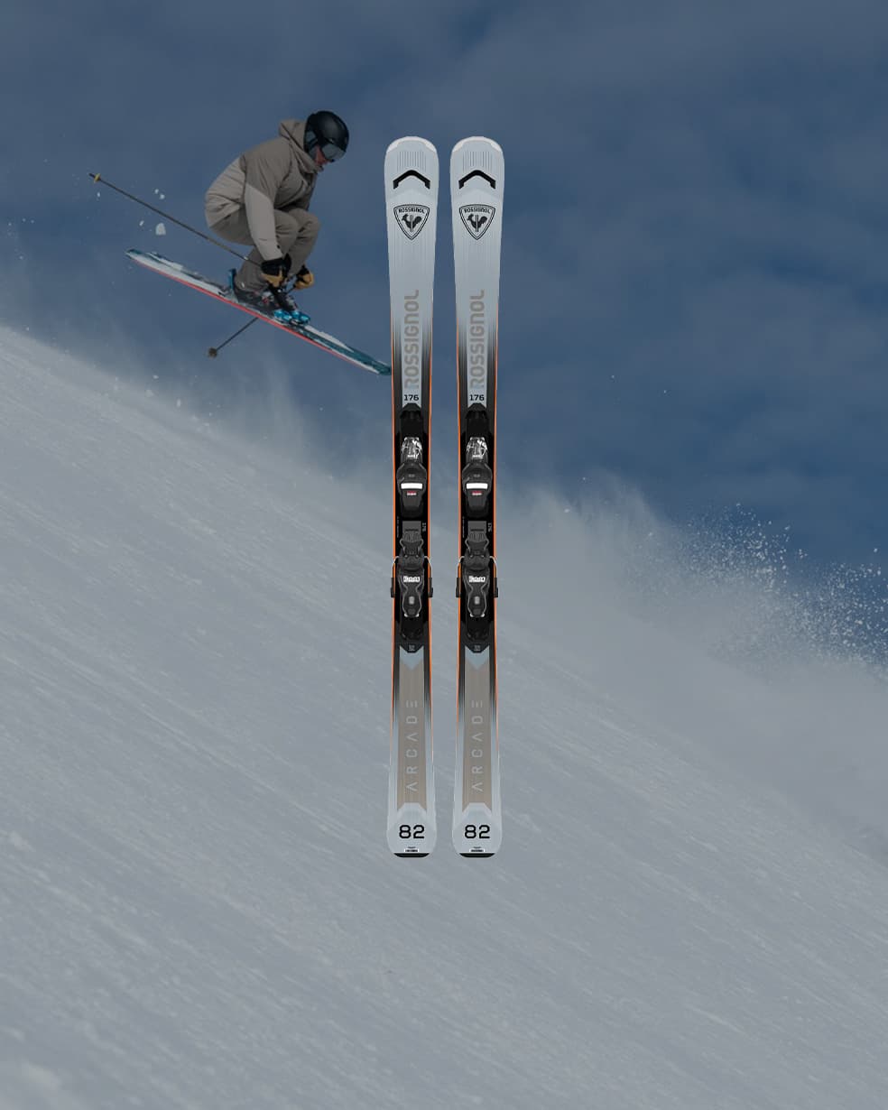 Pair of Rossignol alpine skis centered over a snowy slope while a skier jumps in the background under a cloudy blue sky.