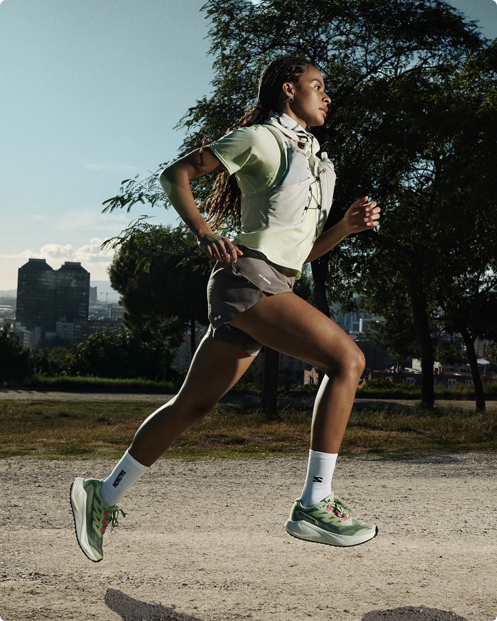 Woman running on a gravel path in a park, wearing a green top, shorts, and running shoes, with trees and city buildings in the background.