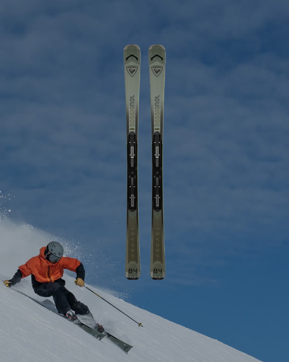 Two alpine skis centered against a blue sky, with a skier in an orange jacket carving down a snowy slope at the lower left.