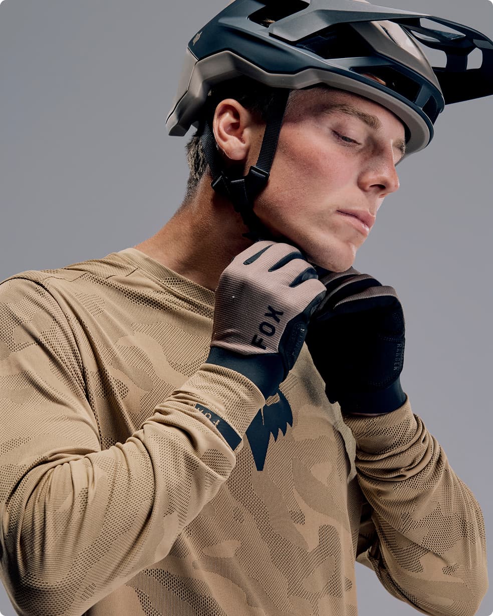 Person fastening a helmet strap, wearing a tan cycling jersey and gloves, against a neutral background.